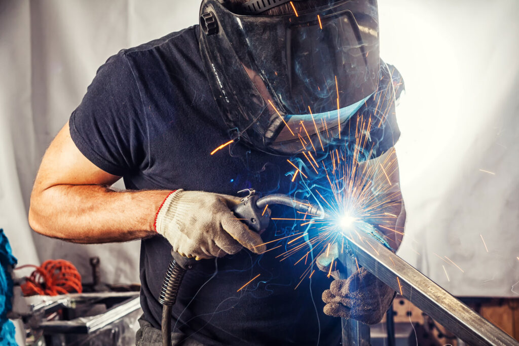 A welder in a black t-shirt and welding helmet with a protective face shield is welding a metal piece, creating bright sparks. The worker wears safety gloves and is focused on the task at hand, demonstrating precision metalwork in a workshop environment.