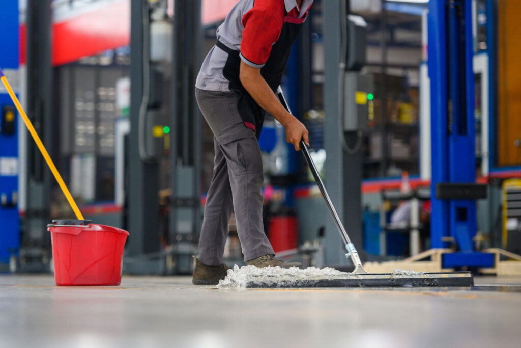 A worker in a gray uniform and safety boots mops the epoxy floor with a wide mop in an industrial setting, with a red bucket and yellow mop handle visible. The background shows the blurred interior of a factory with machinery and blue elements, suggesting a maintenance activity during a work shift.