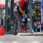 A worker in a gray uniform and safety boots mops the epoxy floor with a wide mop in an industrial setting, with a red bucket and yellow mop handle visible. The background shows the blurred interior of a factory with machinery and blue elements, suggesting a maintenance activity during a work shift.
