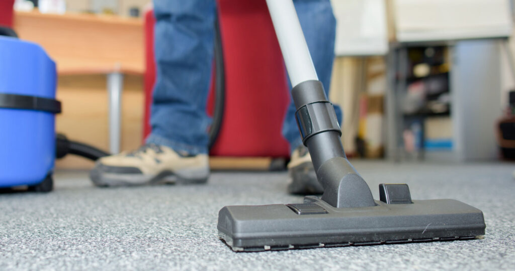 A close-up view of a vacuum cleaner's nozzle on a textured office carpet, with the legs of a person operating it in the background, implying routine cleaning or maintenance work being done in an office environment.