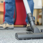 A close-up view of a vacuum cleaner's nozzle on a textured office carpet, with the legs of a person operating it in the background, implying routine cleaning or maintenance work being done in an office environment.