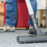 A close-up view of a vacuum cleaner's nozzle on a textured office carpet, with the legs of a person operating it in the background, implying routine cleaning or maintenance work being done in an office environment.
