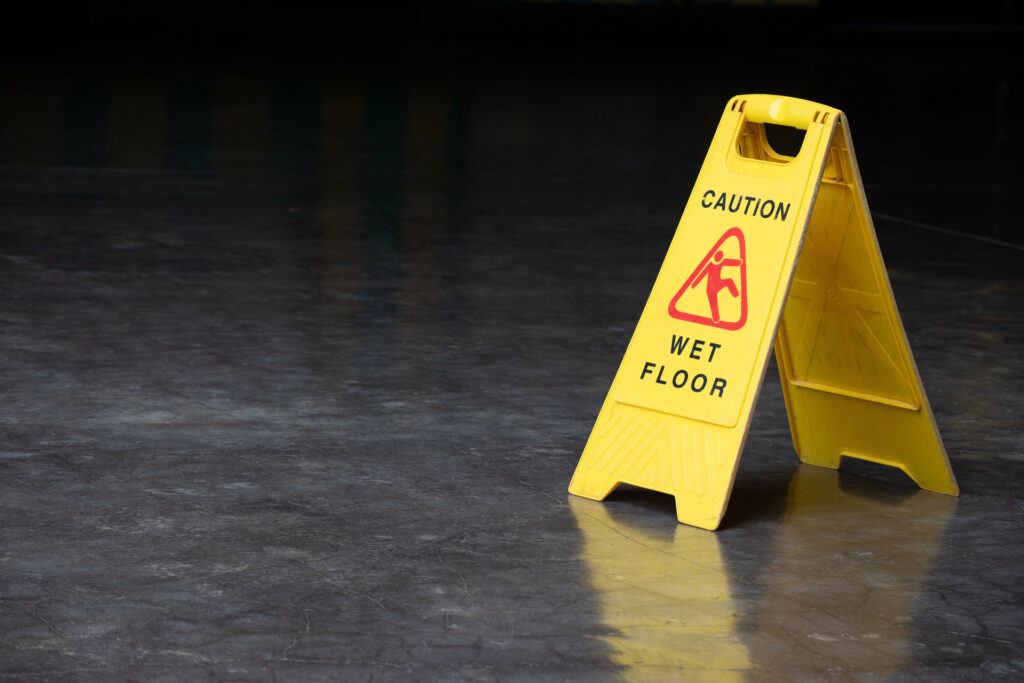 Yellow caution sign on a shiny wet polished concrete floor with the warning 'Wet Floor' and the figure of a person slipping, indicating a hazard and advising people to be careful while walking in the area.