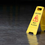 Yellow caution sign on a shiny wet polished concrete floor with the warning 'Wet Floor' and the figure of a person slipping, indicating a hazard and advising people to be careful while walking in the area.