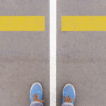 First-person view of a person's feet standing on a gray surface with two large yellow arrows pointing in opposite directions, symbolizing a crossroad or decision-making point.