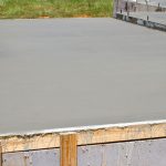 A concrete foundation for a building under construction, with exposed steel rebar and wooden formwork still in place around the edges, set against a backdrop of a grassy area.