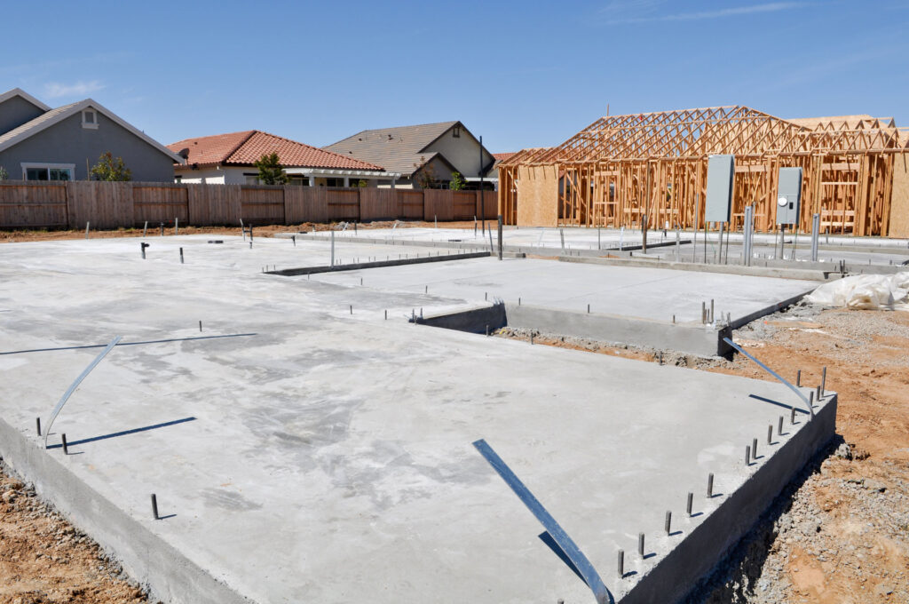 Construction site of a new residential development with the new concrete foundation freshly laid. Steel rebar sticks up from the cement pads, prepared for vertical building. In the background, wooden frameworks of houses under construction are visible, with a clear blue sky above.