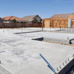 Construction site of a new residential development with the new concrete foundation freshly laid. Steel rebar sticks up from the cement pads, prepared for vertical building. In the background, wooden frameworks of houses under construction are visible, with a clear blue sky above.