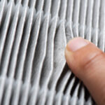 Close-up image of a human finger pressing against a pleated air filter, highlighting the texture and layers of the filtration material.