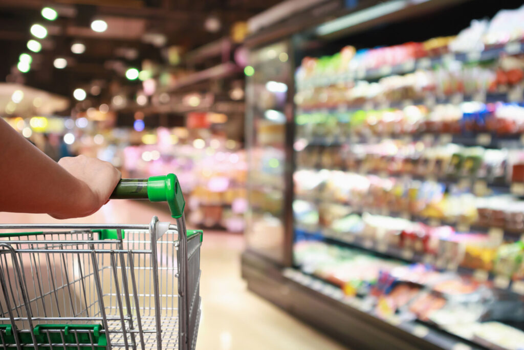 Close-up view of a person's hand holding a green shopping cart handle in a brightly lit supermarket with blurred aisles of products in the background, suggesting the perspective of a shopper.