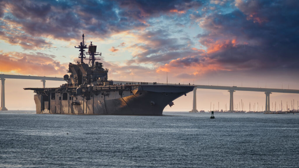 A majestic navy ship at sea during sunset, with a dramatic sky painted with hues of orange, pink, and blue. In the background, a long bridge spans across the horizon, and the calm ocean waters reflect the colors of the sky.