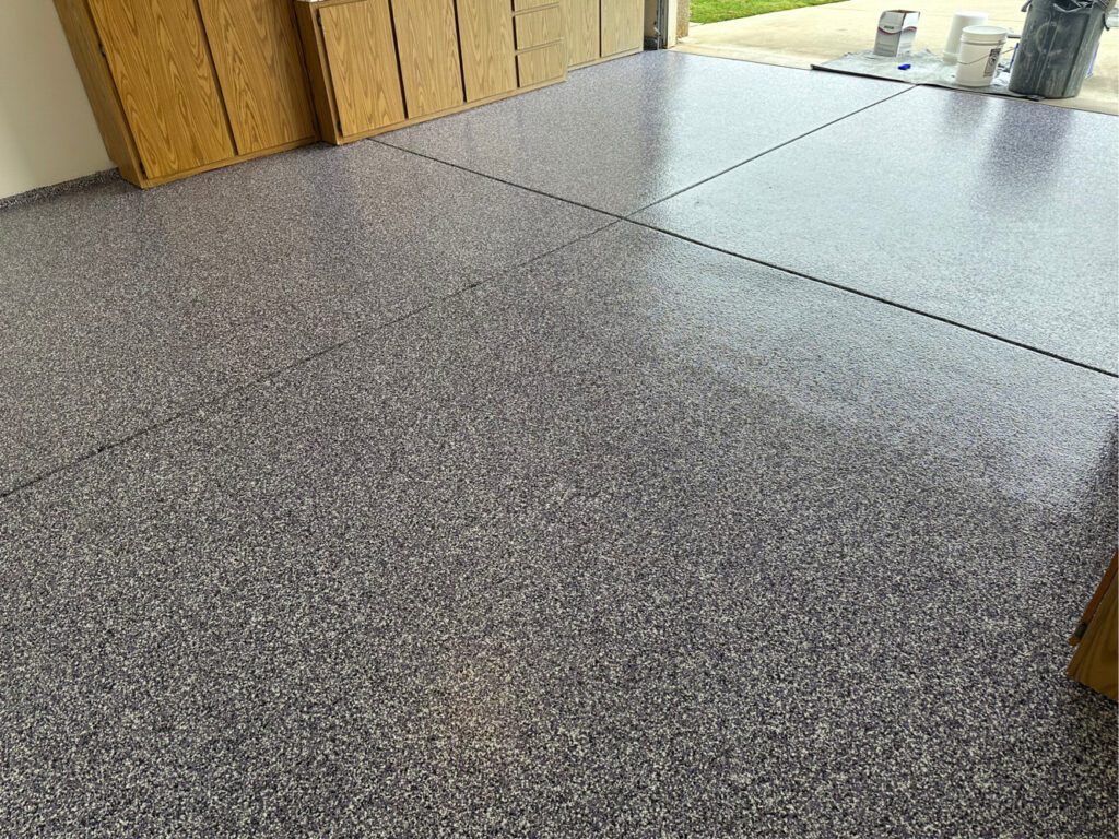 Garage with newly resurfaced epoxy concrete coating. The garage door is open allowing sunlight to pour into this newly refurbished space. The epoxy flooring is flecked with purple and black.