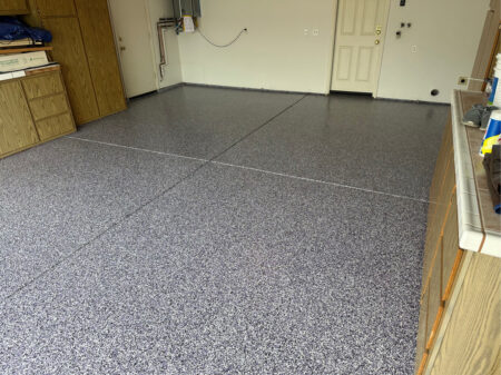 Garage with newly resurfaced epoxy concrete coating. There is a storage cabinet on the left side of the garage and a door to the inside of the home at the back. The epoxy flooring is flecked with purple and black.