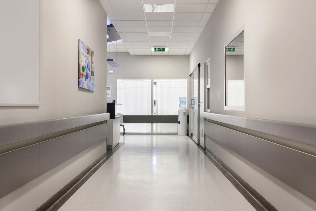 A clean, well-lit hospital hallway with a polished white coved floor and light grey walls. The corridor features handrails on both sides and a few doors leading to various rooms. At the end of the hallway, a water cooler and a small desk are visible near a set of double doors with white curtains. On the left wall, there is a colorful medical-themed artwork depicting healthcare professionals. The ceiling has recessed lighting and an emergency exit sign is clearly displayed. The environment is sterile and orderly, typical of a healthcare facility.