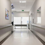 A clean, well-lit hospital hallway with a polished white coved floor and light grey walls. The corridor features handrails on both sides and a few doors leading to various rooms. At the end of the hallway, a water cooler and a small desk are visible near a set of double doors with white curtains. On the left wall, there is a colorful medical-themed artwork depicting healthcare professionals. The ceiling has recessed lighting and an emergency exit sign is clearly displayed. The environment is sterile and orderly, typical of a healthcare facility.