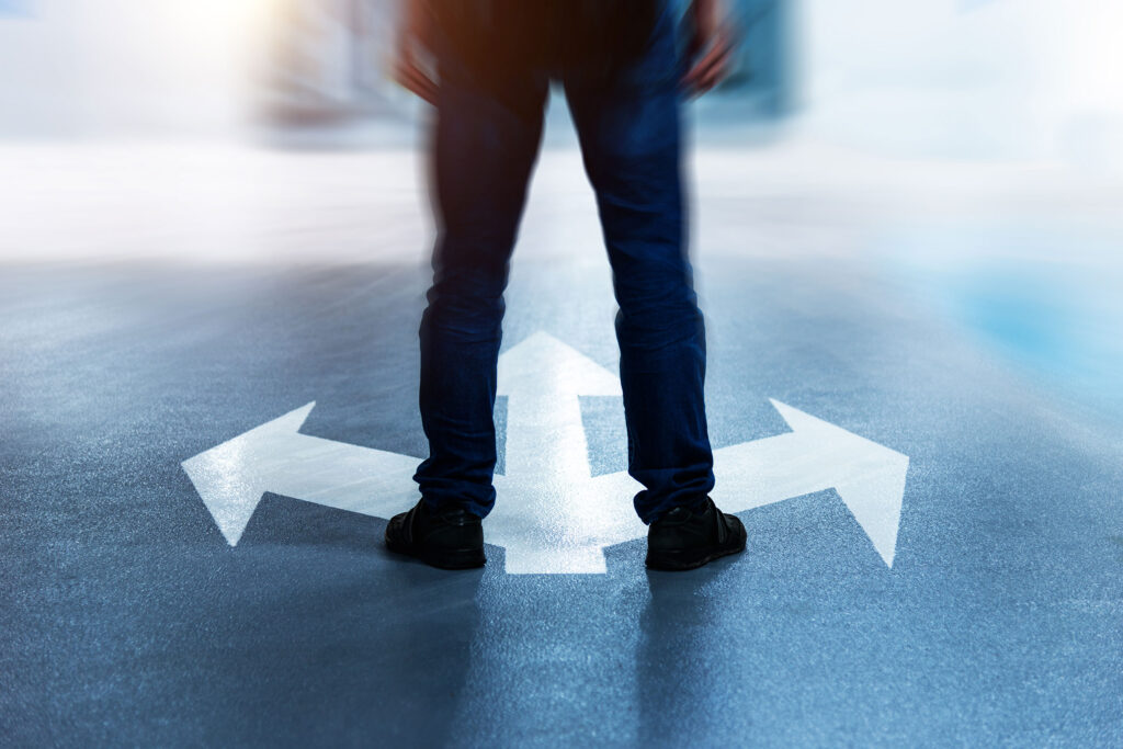 A person stands on a paved surface with three large white arrows painted on the ground, pointing in different directions: left, right, and straight ahead. The figure's legs are visible from the waist down, wearing jeans and black shoes. The image is slightly blurred around the edges, emphasizing the idea of decision-making or choosing a path. The scene is lit with a soft glow, adding to the feeling of contemplation or uncertainty.