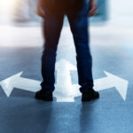 A person stands on a paved surface with three large white arrows painted on the ground, pointing in different directions: left, right, and straight ahead. The figure's legs are visible from the waist down, wearing jeans and black shoes. The image is slightly blurred around the edges, emphasizing the idea of decision-making or choosing a path. The scene is lit with a soft glow, adding to the feeling of contemplation or uncertainty.