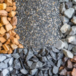 A top-down view of four different types of rocks and gravel, neatly arranged in sections. Clockwise from the top left: coarse orange-brown rocks, small multicolored gravel, grayish-white rocks, and dark brown rocks with a polished appearance. Each type of material contrasts in texture and color, showcasing their distinct characteristics.