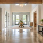 Spacious modern entryway with polished concrete floors and high ceilings. A rustic wooden table with a white orchid centerpiece sits on a textured beige rug in front of large windows with black trim, offering a view of greenery outside. On the left, a large framed botanical artwork hangs on white shiplap walls. On the right, a wooden console table with black metal legs holds a potted fern and decorative items, with a large wooden panel mounted above it. A glass chandelier hangs from the ceiling.