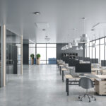 Modern open-plan office with rows of wooden desks equipped with desktop computers and gray ergonomic chairs on a polished concrete floor. Large floor-to-ceiling windows line the right wall, offering views of city skyscrapers. The ceiling features multiple white pendant lights, and two potted plants stand near the windows at the back. Glass-walled private offices or meeting rooms are on the left side. The space has a sleek, minimalistic design with neutral gray and natural wood tones.