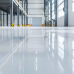 Interior of a modern, empty industrial warehouse with a glossy epoxy-coated floor reflecting the ceiling lights and large windows. The space features high ceilings, steel support beams with yellow and black safety guards, and a large closed roller door at the far end. Natural light fills the area, emphasizing its clean and organized appearance.