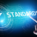 Close-up of a hand pointing toward the word "STANDARDS" on a digital interface with futuristic blue HUD (heads-up display) elements, including gears and circuit-like graphics. A large white checkmark is shown inside a glowing circle, symbolizing approval or compliance.