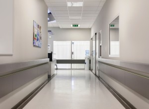 A clean, well-lit hospital hallway with a polished white coved floor and light grey walls. The corridor features handrails on both sides and a few doors leading to various rooms. At the end of the hallway, a water cooler and a small desk are visible near a set of double doors with white curtains. On the left wall, there is a colorful medical-themed artwork depicting healthcare professionals. The ceiling has recessed lighting and an emergency exit sign is clearly displayed. The environment is sterile and orderly, typical of a healthcare facility.