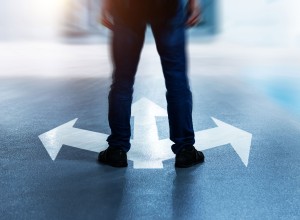 A person stands on a paved surface with three large white arrows painted on the ground, pointing in different directions: left, right, and straight ahead. The figure's legs are visible from the waist down, wearing jeans and black shoes. The image is slightly blurred around the edges, emphasizing the idea of decision-making or choosing a path. The scene is lit with a soft glow, adding to the feeling of contemplation or uncertainty.