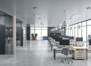 Modern open-plan office with rows of wooden desks equipped with desktop computers and gray ergonomic chairs on a polished concrete floor. Large floor-to-ceiling windows line the right wall, offering views of city skyscrapers. The ceiling features multiple white pendant lights, and two potted plants stand near the windows at the back. Glass-walled private offices or meeting rooms are on the left side. The space has a sleek, minimalistic design with neutral gray and natural wood tones.