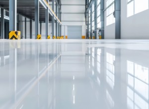Interior of a modern, empty industrial warehouse with a glossy epoxy-coated floor reflecting the ceiling lights and large windows. The space features high ceilings, steel support beams with yellow and black safety guards, and a large closed roller door at the far end. Natural light fills the area, emphasizing its clean and organized appearance.
