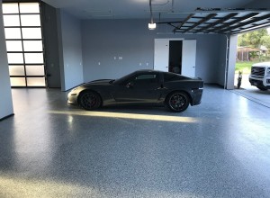 A spacious garage with a speckled grey epoxy floor, housing a sleek dark sports car with red brake calipers. Natural light streams in through the frosted glass of the garage door, casting a warm glow on the vehicle and floor. The clean and organized space conveys a modern look with a touch of luxury, emphasized by the high-end car parked inside.