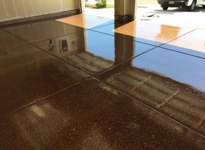 The image showcases a polished, sparkling brown decorative concrete epoxy floor of a garage with a high-gloss finish that reflects the surrounding walls and objects, including a vehicle parked outside. The open garage door lets in natural light, which enhances the reflective quality of the floor, showcasing its pristine condition and the detailed texture of the speckled surface.