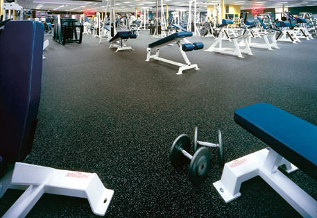 Interior of a modern gym with a variety of exercise equipment in the background and a focus on the black speckled rubber flooring. In the foreground, a weight bench and a loose dumbbell are visible, illustrating a typical gym setting