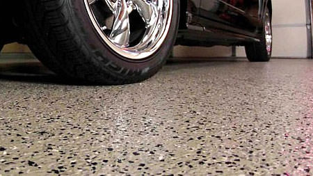Close-up view of speckled epoxy garage flooring with a glossy finish, partially obscured by the tire and chrome wheel of a car parked on it, highlighting the durability and aesthetic of the floor surface in a garage setting.
