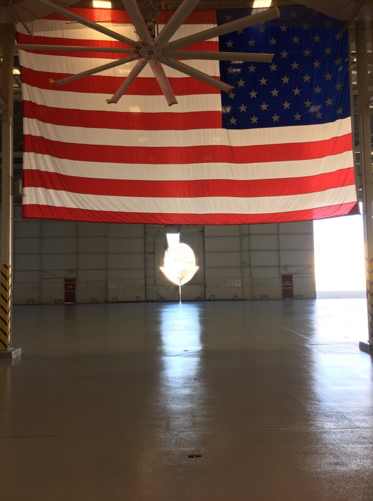 Large American flag hanging from the ceiling in a spacious industrial hangar, with a big ceiling fan above and sunlight streaming in from a doorway, casting a bright reflection on the glossy concrete floor.