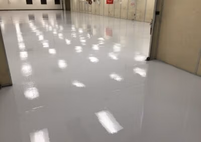 A large industrial or commercial space with a shiny gray floor reflecting the ceiling lights. The periphery of the room features beige walls with several doors and fire extinguisher stations, indicative of a focus on safety. The space appears clean, well-maintained, and possibly used for storage or as a work area.