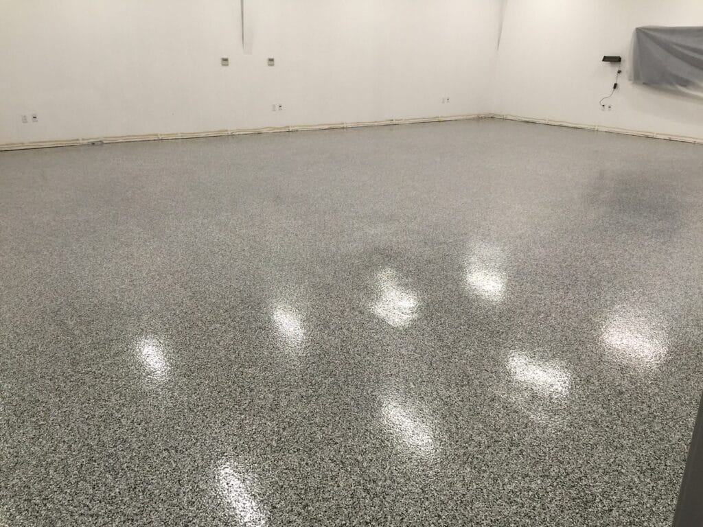 A spacious room with a speckled grey epoxy floor that reflects overhead lighting. White walls with electrical outlets and a baseboard run around the perimeter. A plastic sheet partially covers a segment of the wall on the right side, suggesting an area under construction or renovation.