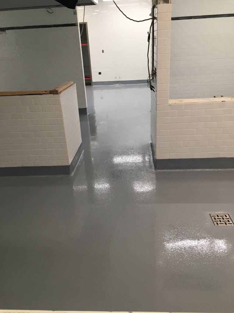 Elevated view of a room with a light gray epoxy floor that glistens under the lighting, with white tiled walls and exposed electrical conduits. A section of the wall features red safety markings near a floor drain, and the space appears to be part of an industrial or commercial setting.