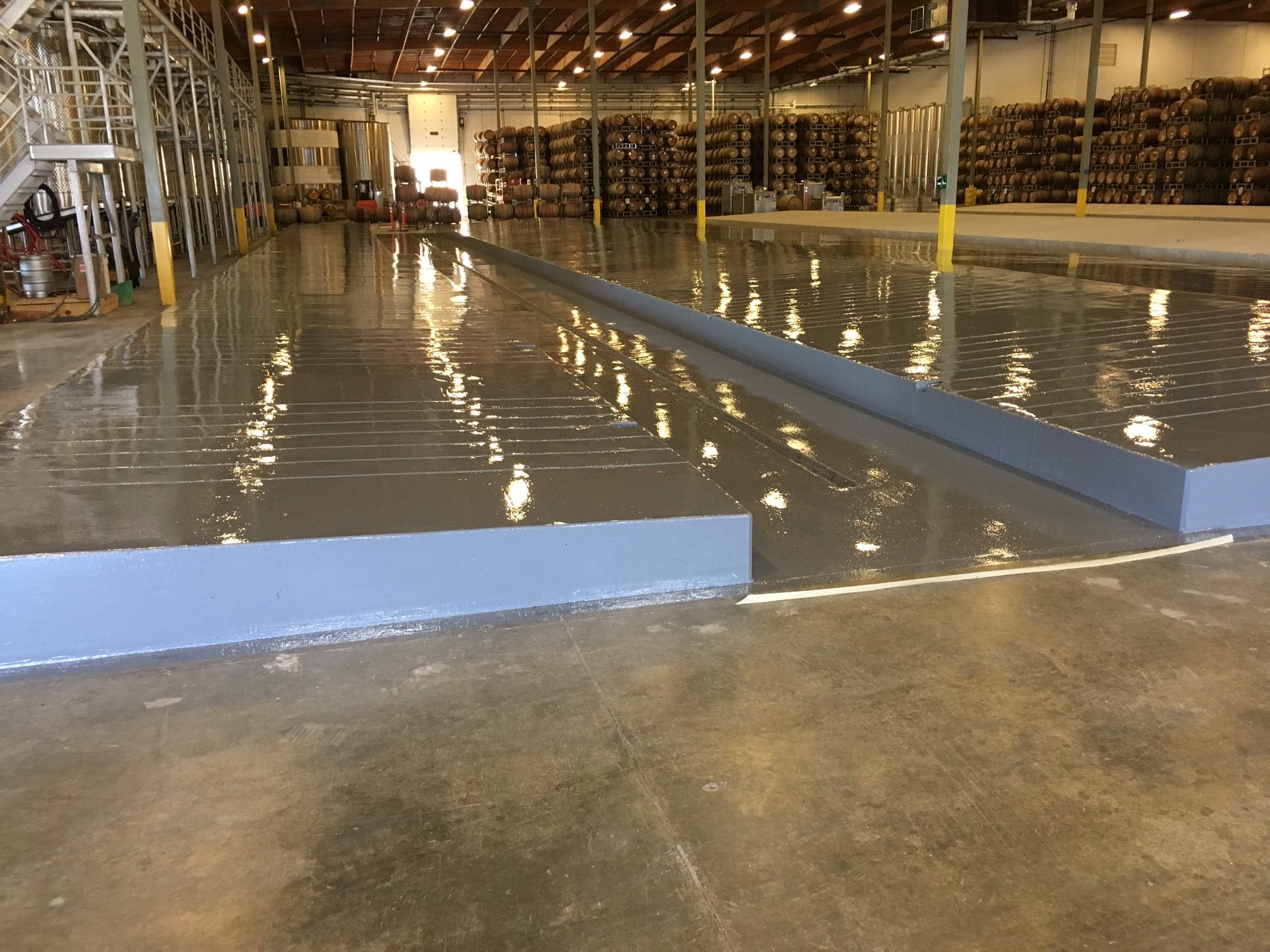 Natural Sealed Concrete in Warehouse Large industrial warehouse with a glossy epoxy floor coating that reflects the bright ceiling lights. A prominent, long blue ramp runs through the space, providing access between different floor levels. Rows of stocked shelves with goods on pallets are visible in the background, highlighting the warehouse's storage function.