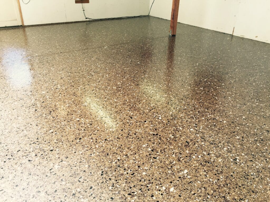 A room with a high-gloss epoxy floor, densely speckled in shades of brown, beige, and black. The light reflects off the smooth surface creating a warm glow. In the background, there is a simple wooden post and white walls with some scuff marks, indicating the space is used for practical purposes, possibly a workshop or a garage.