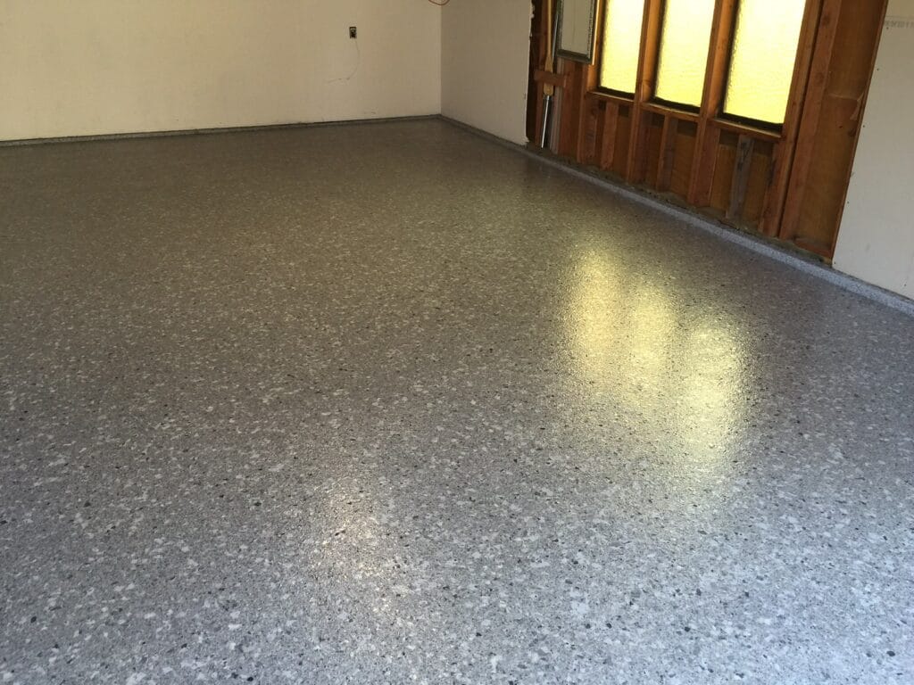 A room with a gray speckled epoxy floor casting a light sheen. Sunlight streams in through textured glass windows set in a wooden frame on the right wall, creating bright patterns on the floor. The left wall is white with a visible electrical outlet and a bare corner. This image suggests a residential space with a blend of warmth and functionality.