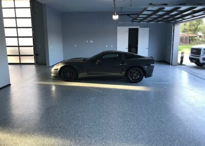A spacious garage with a speckled grey epoxy floor with top coat, housing a sleek dark sports car with red brake calipers. Natural light streams in through the frosted glass of the garage door, casting a warm glow on the vehicle and floor. The clean and organized space conveys a modern look with a touch of luxury, emphasized by the high-end car parked inside.