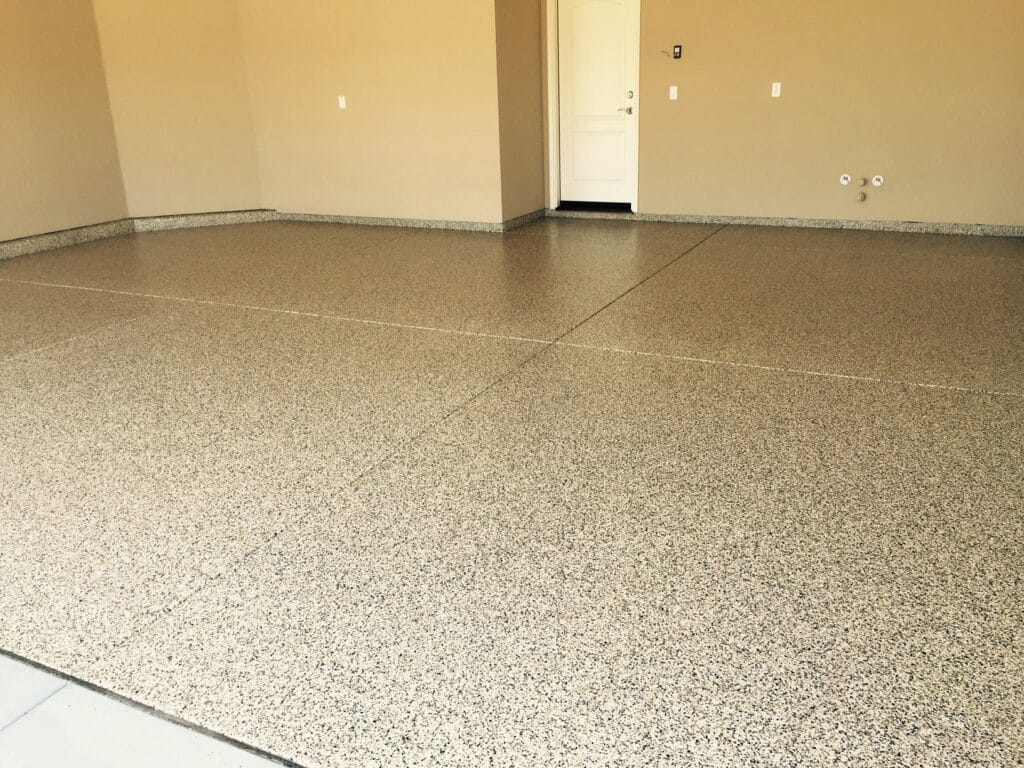 An empty room with speckled terrazzo flooring and beige walls. There is a white door on the far wall with a light switch to its right and two electrical outlets on adjacent walls. The room has a baseboard that matches the floor, providing a seamless transition between the wall and floor.