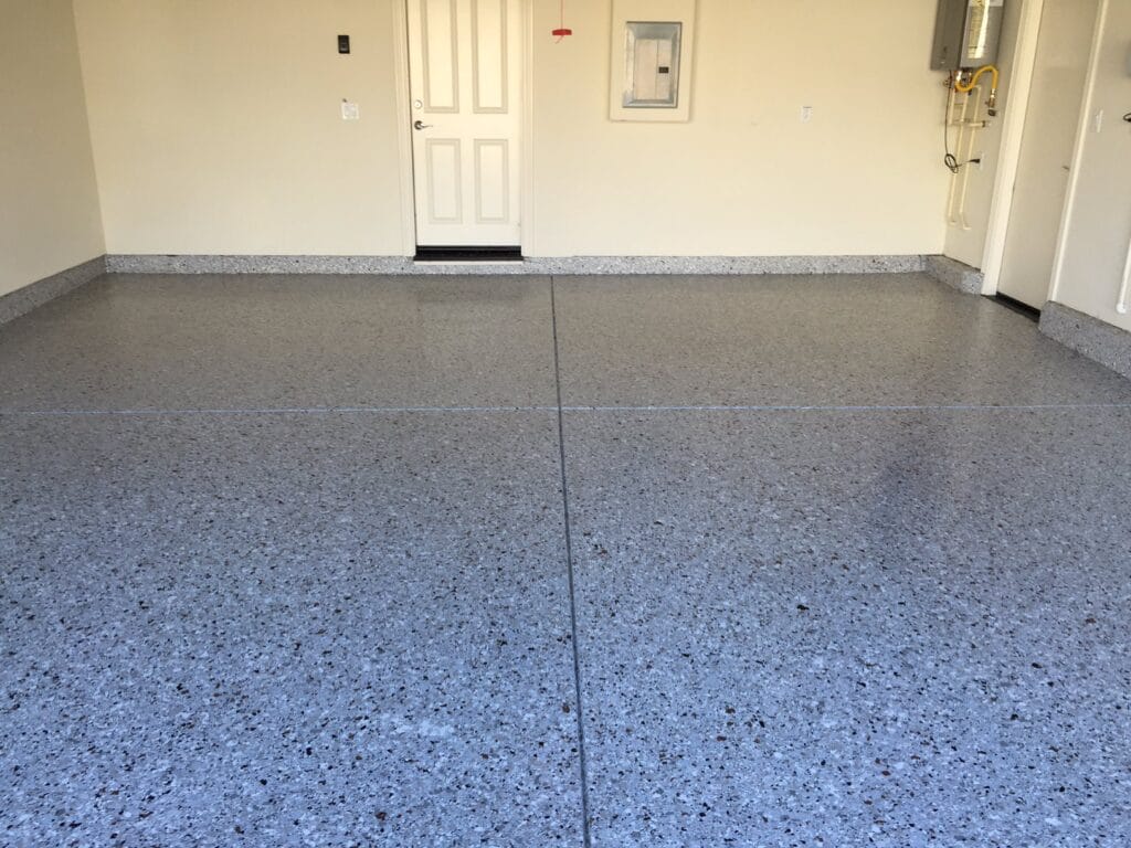 A clean, empty room with a light gray speckled epoxy floor. There are two white doors on the back wall, one closed and the other slightly open. To the right, there's a water heater and gas pipeline. The room also features a small white electrical panel on the right wall. The space looks like a functional area, possibly a laundry or utility room in a residential home.