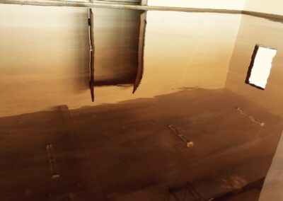 A large, empty room with a high-shine caramel-colored epoxy floor reflecting the ceiling's structure and a bright window. The reflections create a mirrored effect, giving the impression of a vast and open space. The smooth surface of the floor and its reflective quality indicate a modern and well-maintained interior, possibly in a commercial or industrial setting.