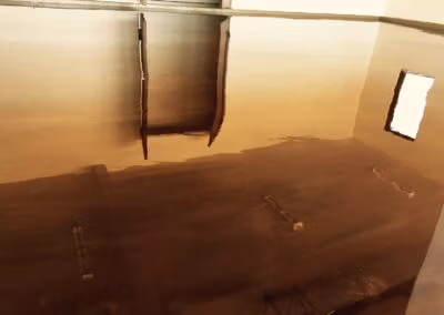 A large, empty room with a high-shine caramel-colored epoxy floor reflecting the ceiling's structure and a bright window. The reflections create a mirrored effect, giving the impression of a vast and open space. The smooth surface of the floor and its reflective quality indicate a modern and well-maintained interior, possibly in a commercial or industrial setting.