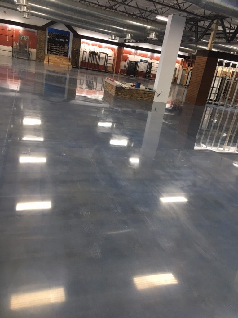 An expansive, well-lit interior space with a high ceiling and reflective polished concrete floor. The area has a variety of fixtures, including a reception desk with a stone facade, red and wood-toned wall accents, and metal-framed glass partitions. Ceiling-mounted lights cast a series of bright reflections on the floor. It looks like a commercial setting, possibly a lobby or retail environment, with some areas cordoned off, indicating potential construction or rearrangement.