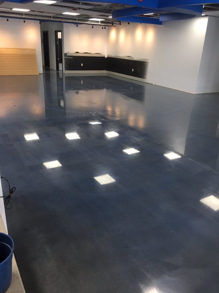 A large room with a polished blue-gray epoxy floor that reflects the ceiling's recessed lights. The space features a mix of white and black walls, with a black counter or partition area towards the back. Blue accents are seen overhead, possibly part of the ceiling design. The room is empty except for a blue bucket in the foreground, suggesting a cleaning process or recent maintenance. The ambiance is modern and appears to be part of a commercial or public facility.