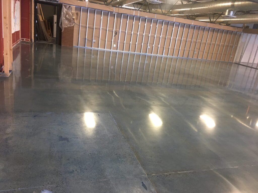 Spacious indoor area with a concrete floor reflecting overhead lights, creating a glossy surface. To the right, a wooden railing partitions the space, and beyond it, metal ductwork is visible against the ceiling. An open doorway on the left reveals a glimpse into another room with visible construction materials. The photo conveys a sense of ongoing interior work or renovation in a commercial or industrial building.