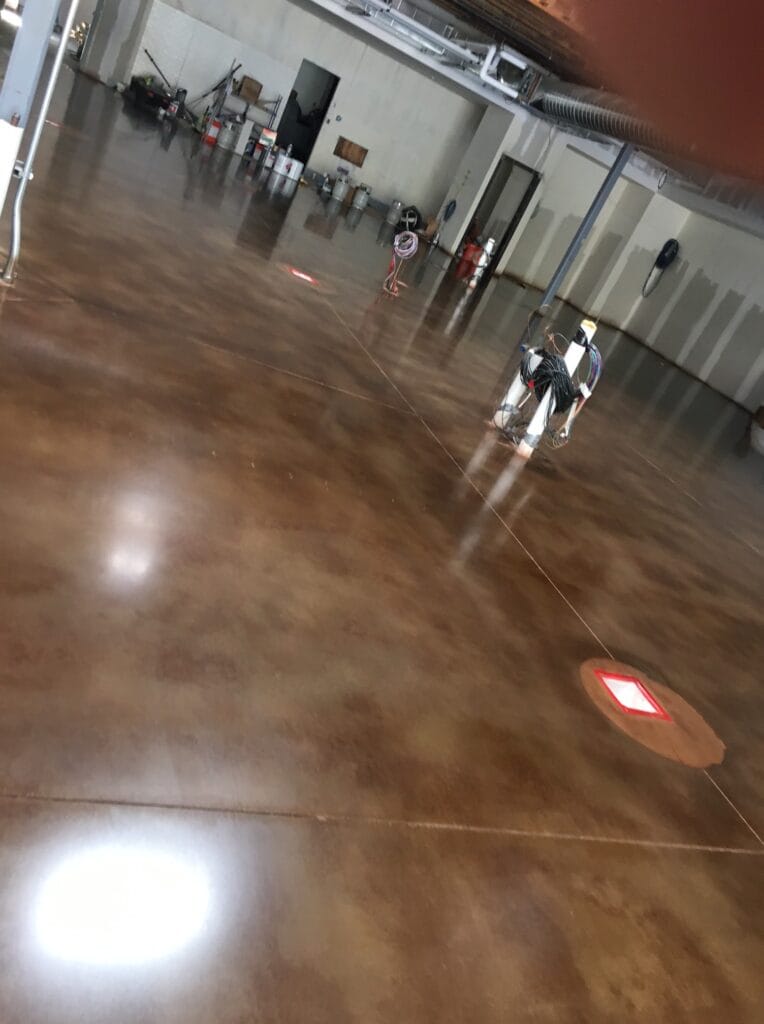 An indoor space with glossy polished concrete flooring, showing a tilted perspective. A person in a wheelchair is seen in the distance, centered and facing away, with cables draped over the back. The room features industrial elements like exposed pipes and a metal door, with cleaning equipment stored along the wall. A reflective red floor sign is visible closer to the foreground, slightly to the left.